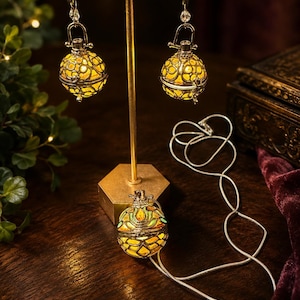 May include: A set of jewelry featuring two spherical, filigree-style earrings and a matching pendant on a silver-toned chain. Each piece has a glowing yellow interior visible through the intricate metal lattice design, suspended from a gold-toned stand.