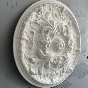 Op de afbeelding: Een cirkelvormig wit plafondmedaillon met een ingewikkeld reliëfpatroon van acanthusbladeren, krullende ranken en een centrale geribbelde koepel. Het decoratieve architecturale element heeft een verhoogde rand met een herhalend ribbelpatroon.