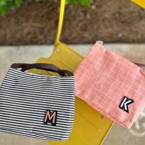 May include: Two lunch bags with personalized initials. The bag on the left is black and white striped with the letter "M" on the front. The bag on the right is pink with a grid pattern and the letter "K" on the front.