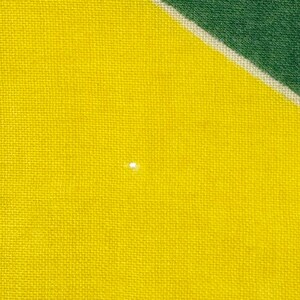 Pode incluir: Close-up de um tecido amarelo com uma trama texturizada. Uma pequena mancha branca brilhante &eacute; vis&iacute;vel na superf&iacute;cie amarela. O canto superior direito apresenta uma se&ccedil;&atilde;o diagonal de tecido verde, separada por uma faixa branca.