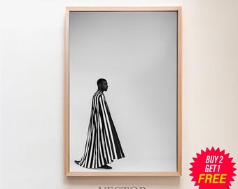 Puede incluir: Fotografía en blanco y negro de una persona con una capa a rayas, de pie en una habitación blanca. La imagen está enmarcada en un marco de madera claro.