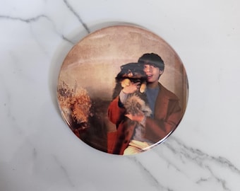 May include: A round button featuring a vintage-style image of a person holding a small dog. The person is wearing a brown coat over a blue shirt. The dog has black and brown fur. The button has a glossy finish.