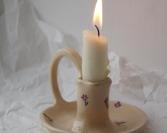 May include: A ceramic candle holder with a floral design and a single white candle burning inside. The holder has a handle and a small base.