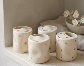 May include: Four off-white ceramic containers with a wavy top and a small opening. The containers are decorated with pink flowers, blue stars, and red hearts. They are arranged on a light-colored shelf.