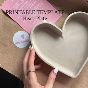 Puede incluir: Un plato de cerámica blanco en forma de corazón con un borde ligeramente elevado. El plato está sobre una superficie marrón. El texto "PRINTABLE TEMPLATE Heart Plate" es visible en el fondo.