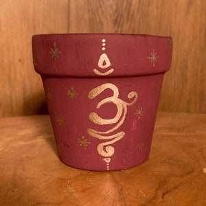 May include: A small terracotta flower pot painted in a deep burgundy red with gold accents. The pot features a gold Om symbol and a pattern of gold snowflakes.