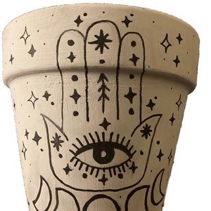 Puede incluir: Una maceta de cerámica blanca con un diseño de mano negra, un ojo y lunas. El diseño de la mano incluye estrellas y otros símbolos.