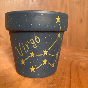 Puede incluir: Una maceta de terracota azul oscuro con un diseño de constelación y signo del zodíaco Virgo dorado.