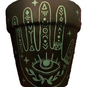 May include: Black terracotta planter pot with a hand painted design of a hand with an eye in the palm. The hand is outlined in teal and has various geometric patterns on the fingers. The eye is also teal with black details.