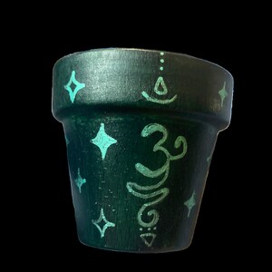 May include: A small, dark green painted flower pot with turquoise accents. The pot features a hand-painted Om symbol and star-shaped designs. The pot is designed for small plants and adds a touch of bohemian style.
