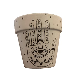 May include: White ceramic planter with a black hand design featuring an eye and geometric patterns. The hand is drawn in a stylized, almost tribal style.
