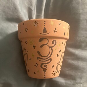 May include: A terracotta planter with a light pink glaze and gold and black painted designs. The design includes a symbol that looks like a stylized "3" and other geometric shapes.