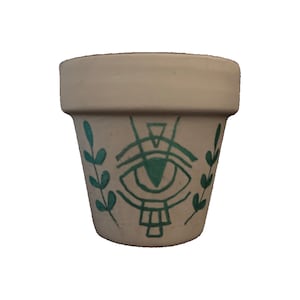 Puede incluir: Una maceta pequeña de terracota blanca con un diseño de ojo verde y ramas con hojas a cada lado. El ojo está rodeado de un círculo y tiene un triángulo encima.