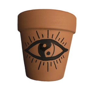 Puede incluir: Maceta de terracota con un símbolo yin yang en blanco y negro dentro de un diseño de ojo.