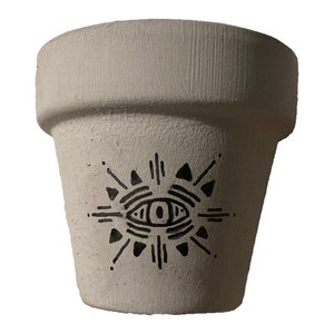Puede incluir: Maceta de cerámica blanca con un diseño de ojo en blanco y negro. El ojo está rodeado de un patrón de rayos de sol.