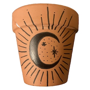 Puede incluir: Una maceta de terracota con una luna creciente negra y estrellas pintadas en ella. La luna está rodeada por un diseño de rayos de sol.