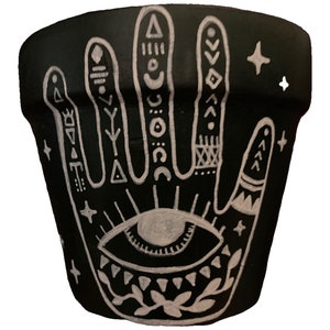 May include: Black terracotta planter with a white hand design featuring an eye, stars, and geometric patterns.