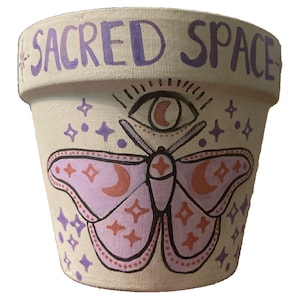May include: A white terracotta pot with the words "SACRED SPACE" painted in purple on the top rim. The pot is decorated with a purple butterfly with orange accents, a crescent moon, and a stylised eye with a crescent moon inside.