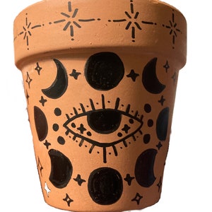 Puede incluir: Maceta de terracota con un diseño pintado en blanco y negro que presenta una luna creciente, estrellas y un ojo.
