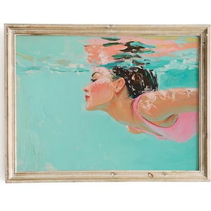 Puede incluir: Una pintura de una mujer nadando bajo el agua con un traje de baño rosa. El agua es de color azul turquesa y el cabello de la mujer es castaño oscuro. La pintura está enmarcada en un marco de madera clara.