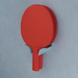 May include: A red table tennis paddle is held in place by a light blue wall-mounted holder. The paddle features a circular head and a cylindrical handle, while the holder has two curved arms that support the neck of the paddle against the wall.