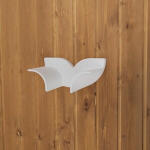 May include: A white wall-mounted shelf featuring a curved, wing-like silhouette with two distinct sections. The shelf has a smooth, matte finish and is designed with sweeping, organic lines that extend outward from the center mounting point.