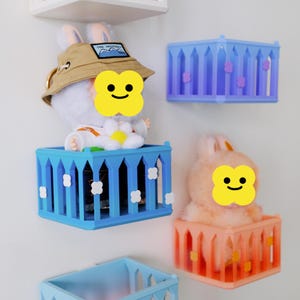 May include: A collection of colorful, decorative wall-mounted cages, each holding a plush bunny. The cages are in shades of blue and orange, with white and purple accents. One bunny wears a hat with text.
