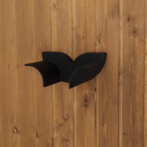 May include: A matte black wall-mounted headphone stand featuring a curved, wing-like design with two distinct cradles for resting headphones. The structure is solid and minimalist, designed to be attached directly to a vertical surface for storage.