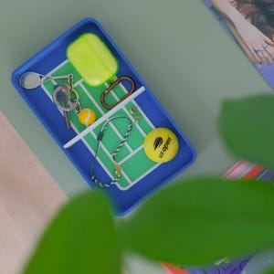 Puede incluir: Una bandeja azul con diseño de cancha de tenis contiene una pelota de tenis amarilla, una funda verde, un llavero con una raqueta de tenis en miniatura y una pulsera. La pelota de tenis tiene la inscripción "US Open".