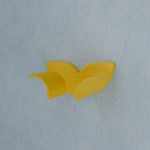 May include: A bright yellow wall-mounted hook featuring a curved, leaf-like design with two distinct, sweeping sections that extend outward. The object has a smooth, matte finish and is attached to a light blue, textured vertical surface.