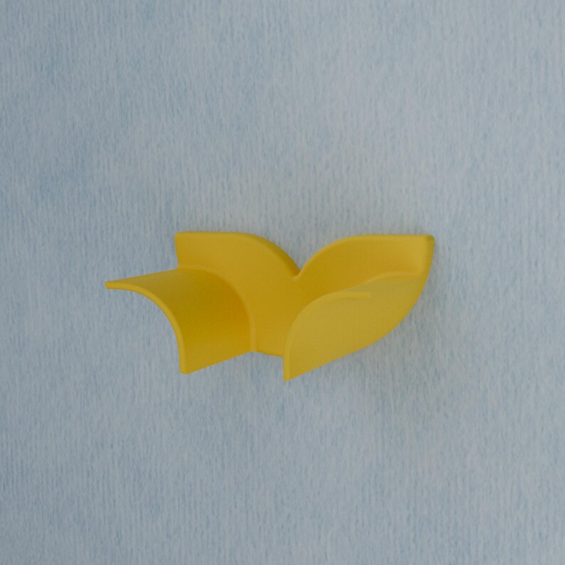 May include: A bright yellow wall-mounted hook featuring a curved, leaf-like design with two distinct, sweeping sections that extend outward. The object has a smooth, matte finish and is attached to a light blue, textured vertical surface.