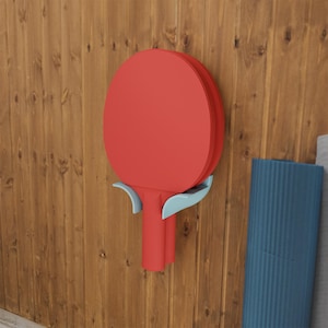 Puede incluir: Una paleta de tenis de mesa roja apoyada en un soporte de pared azul claro. La paleta presenta una cabeza circular y un mango rectangular, colocada de forma segura dentro de los brazos curvos del soporte, fijado a una superficie vertical.