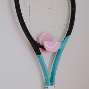 Può includere: Una racchetta da tennis con telaio nero e manico turchese è montata su una parete bianca. Un porta palline da tennis rosa e bianco è attaccato alla racchetta. La racchetta è incordata con corde bianche.