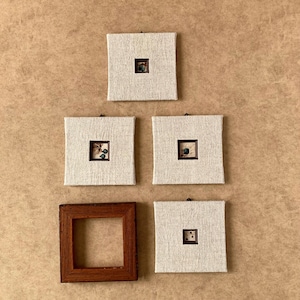 Pode incluir: Cinco peças de arte de parede quadradas. Quatro são quadrados de tecido branco sujo com pequenos quadrados centrais emoldurados escuros contendo elementos decorativos. Uma é uma moldura quadrada de madeira escura. O fundo é marrom claro.