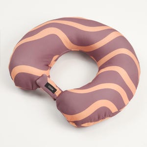 May include: A donut-shaped travel pillow with a wavy pattern in shades of pink and orange. The pillow has a black label with the word "volta" on it.