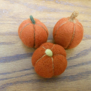 May include: Three felt pumpkins in orange with green and brown stems. The pumpkins are on a wood surface.