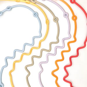 Könnte beinhalten: Ein Set aus sieben bunten Silikonketten mit welligem Design. Jede Kette hat an einem Ende einen kleinen Vogelanhänger und entlang der Länge eine Reihe kleiner Kreise.