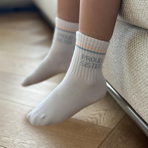 Puede incluir: Un par de calcetines blancos de caña media con puños acanalados que presentan finas rayas horizontales en naranja y azul. El texto PROUD SISTER está impreso en una fuente gris oscuro sin remates en el lado exterior de cada calcetín.