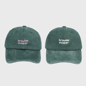 Könnte beinhalten: Zwei dunkelgrüne Baseballkappen mit einer verwaschenen, abgenutzten Textur. Jede Kappe zeigt den gestickten Schriftzug "trouble maker" mittig auf der Vorderseite. Die linke Kappe hat eine rosa Stickerei, die rechte eine weiße Stickerei.