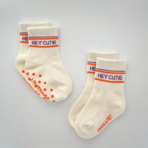 Könnte beinhalten: Cremefarbene Knöchelsocken mit dem Text "HEY CUTIE" in Orange und Lila. Die Socken haben orangefarbene Anti-Rutsch-Punkte auf den Sohlen und den Markennamen "PIPA & PIET" in Orange. Die Socken sind am oberen Rand gerippt.