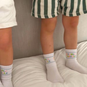 Puede incluir: Dos personas con calcetines blancos con el texto "DOUBLE" en colores. Una persona lleva pantalones cortos de rayas verdes y blancas.
