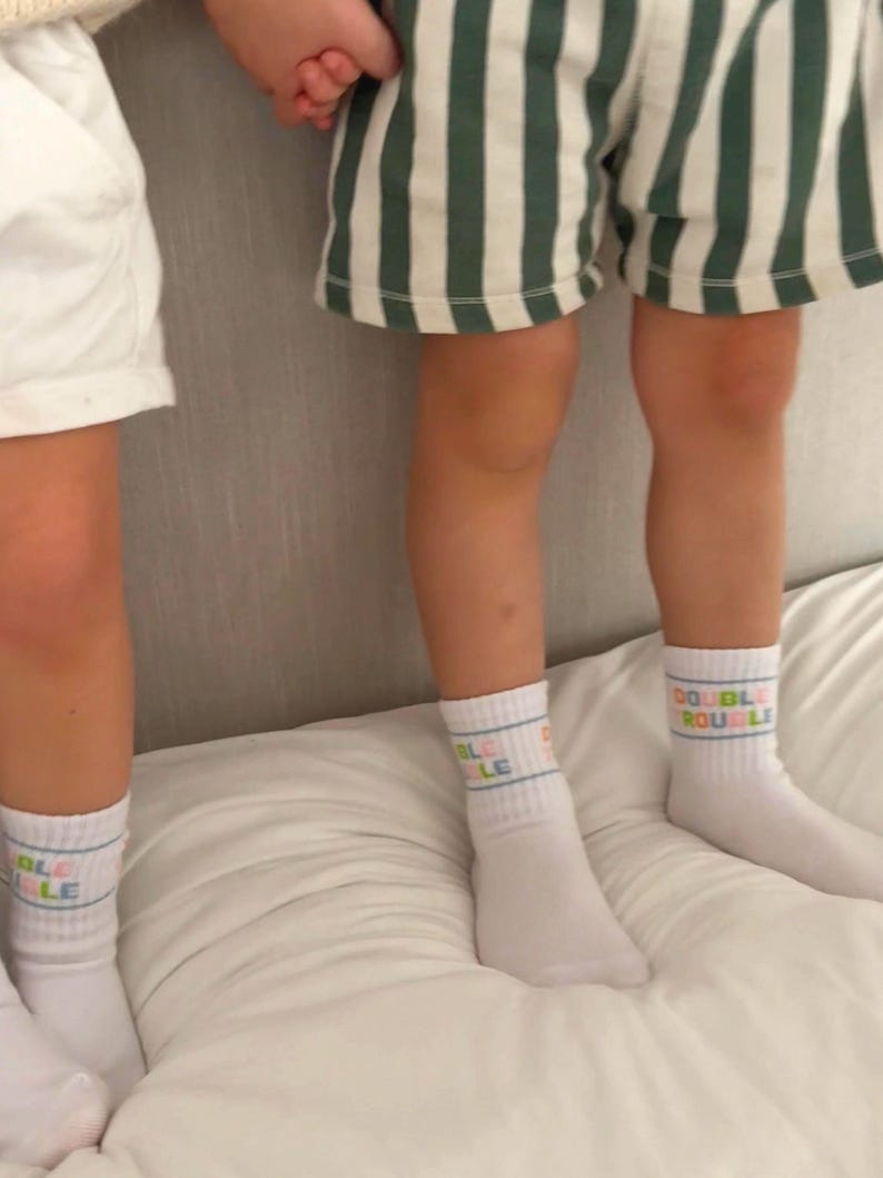 Puede incluir: Dos personas con calcetines blancos con el texto "DOUBLE" en colores. Una persona lleva pantalones cortos de rayas verdes y blancas.