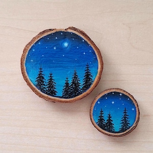 Pode incluir: Duas fatias redondas de madeira com cenas pintadas de um céu noturno azul escuro com estrelas brancas e uma lua crescente, e uma silhueta de uma floresta de pinheiros.