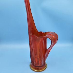 May include: A tall, red glass vase with a unique, curved design. The vase has a wide opening and a narrow base. It is perfect for displaying flowers or other decorative items.