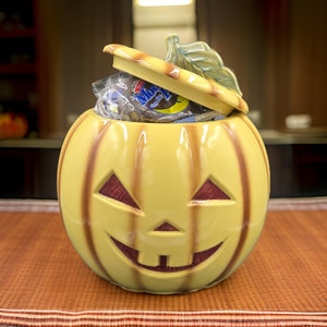 May include: A yellow ceramic pumpkin-shaped candy jar with a brown lid and a carved jack-o'-lantern face. The lid is open and the jar is filled with chocolate candy.