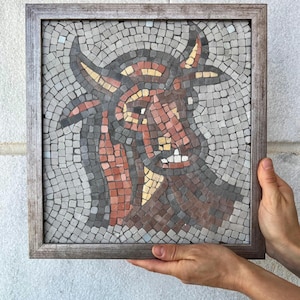 May include: A mosaic artwork depicting a bull's head, framed in a rustic wooden frame. The mosaic is composed of small, square tiles in shades of gray, brown, red, and yellow. The bull's head is centered, with horns and facial features clearly defined.