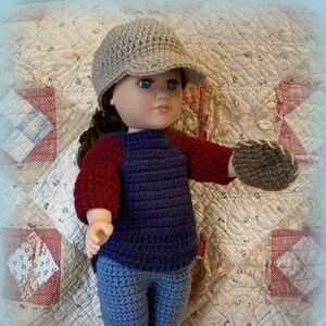 Könnte beinhalten: Eine Puppe, die ein gehäkeltes Outfit trägt, darunter ein blau-rot gestreiftes Shirt, eine blaue Jeans, eine braune Baseballkappe und braune gehäkelte Fäustlinge. Die Puppe steht auf einer weißen und roten Steppdecke.