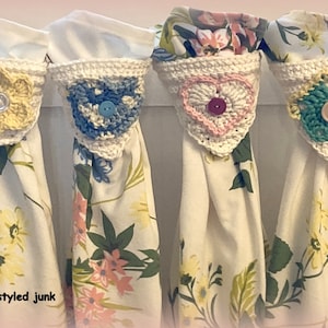 May include: Four white kitchen towels with floral patterns are hanging from a rack. Each towel has a crocheted heart-shaped embellishment with a button in the center. The hearts are in different colors: yellow, blue, pink, and green.