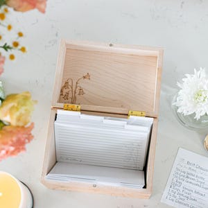 Wooden recipe box sits on a kitchen counter. The lid of the recipe box is open. Over head view of the recipe box shows floral etching on the bottom of the lid and white recipe cards and dividers inside the box.