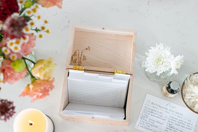 Wooden recipe box sits on a kitchen counter. The lid of the recipe box is open. Over head view of the recipe box shows floral etching on the bottom of the lid and white recipe cards and dividers inside the box.